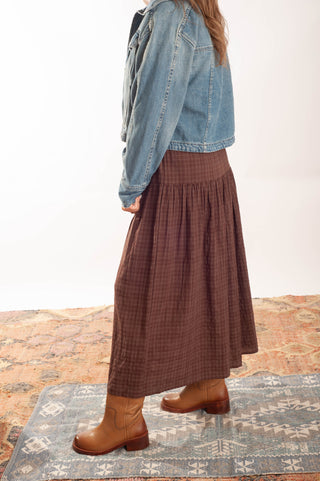 A person shown from the side wearing a brown plaid midi skirt, a light blue denim jacket, and tan boots, standing on a patterned rug against a white studio background.