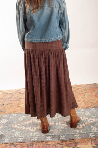 A person shown from the back wearing a brown plaid midi skirt with a wide elastic waistband, paired with a blue denim jacket and brown leather boots, standing on a patterned rug.