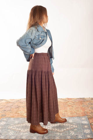A woman stands in profile wearing a brown plaid maxi skirt, a light blue denim jacket, a white top, and brown leather boots, standing on a patterned rug against a white studio background.