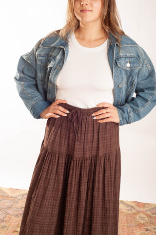 A woman wearing a brown tiered plaid maxi skirt with a drawstring waist, styled with a white ribbed tank top and a light blue denim jacket, standing against a white studio background.