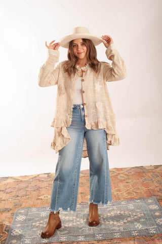 A woman stands in a studio setting wearing a cream-colored textured button-up cardigan with ruffled edges, a white top, wide-leg blue jeans, and brown boots. She is also wearing a large cream-colored cowboy hat, holding it up with both hands.