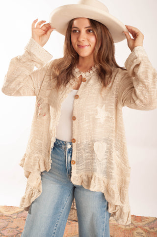 A woman wearing a cream-colored, textured button-up cardigan with star and heart appliques, a white top, blue jeans, and a matching wide-brim hat, posing against a white studio background.