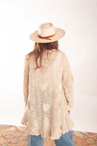 A woman viewed from behind wearing a light beige textured long-sleeve tunic top with heart motifs, blue jeans, and a cream-colored wide-brim hat with a brown band, standing against a plain white wall.