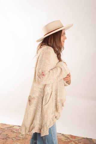 A woman wearing a cream-colored, textured bohemian-style cardigan, a wide-brimmed beige hat, and blue jeans, standing against a plain white studio background.