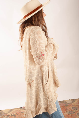 A woman wearing a cream-colored, textured open-front cardigan with floral embroidery on the sleeves and hem, paired with a wide-brimmed cream hat and blue jeans, shown from a side-back angle against a plain white background.