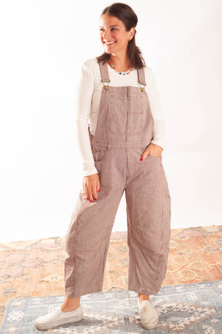 A woman modeling a pair of light brown corduroy overall-style pants over a white long-sleeve top, standing on a patterned rug against a white wall.
