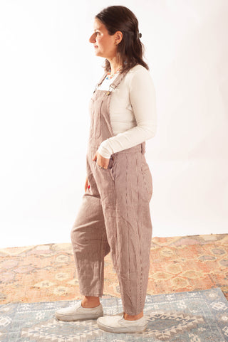A woman wearing a pair of dusty taupe-brown linen overall jumpsuits over a white long-sleeve shirt, standing in profile against a white studio background.