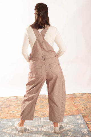 A woman viewed from behind wearing wide-leg brown corduroy overall pants over a white long-sleeve top, standing on a patterned rug against a white studio background.