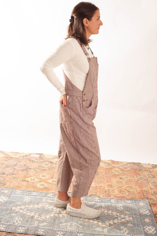 A woman wearing brown corduroy overall pants over a white long-sleeve top, standing in profile on a patterned rug against a white studio background.