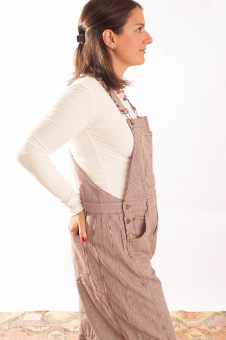 A woman in profile wearing brown corduroy overall-style overalls over a white long-sleeve top, photographed against a plain white studio background.