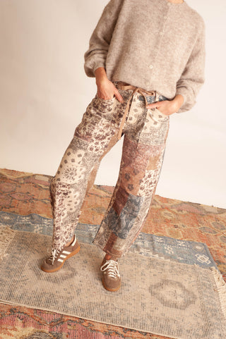 Women modelling paisley printed patchwork pants against a neutral photo backdrop and standing on boho printed rugs. 