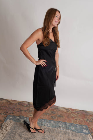 Women wearing a black lace trim sating dress against a neutral photo backdrop standing on boho inspired rugs. 