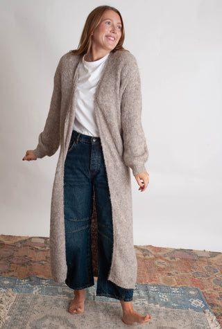 Women modelling a long beige cardigan against a neutral photo backdrop standing on boho inspired printed rugs. 