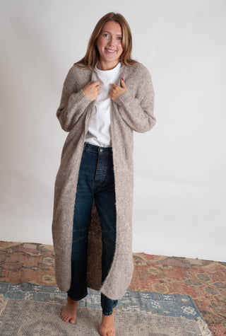 Women modelling a long beige cardigan against a neutral photo backdrop standing on boho inspired printed rugs. 