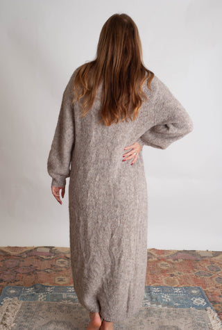 Women modelling a long beige cardigan against a neutral photo backdrop standing on boho inspired printed rugs. 