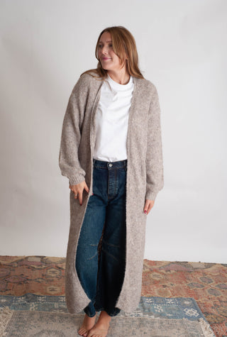 Women modelling a long beige cardigan against a neutral photo backdrop standing on boho inspired printed rugs. 