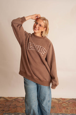 A woman wearing a brown crewneck sweatshirt with 'BLONDE' printed in arched varsity letters across the chest, paired with light blue jeans, standing against a plain beige wall.