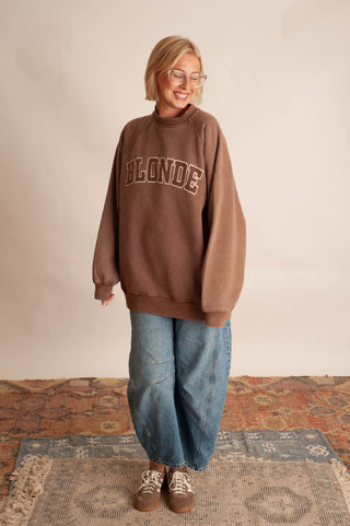 A woman wearing a brown oversized crewneck sweatshirt with 'BLONDE' printed in brown block letters across the chest, paired with wide-leg blue jeans and brown sneakers, standing on a patterned rug against a plain beige wall.