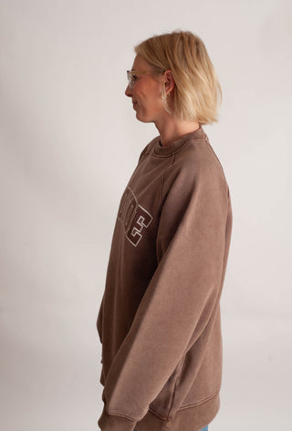 A woman with short blonde hair and glasses, shown in profile wearing a brown crewneck sweatshirt with a white graphic print on the chest, set against a plain light gray background.