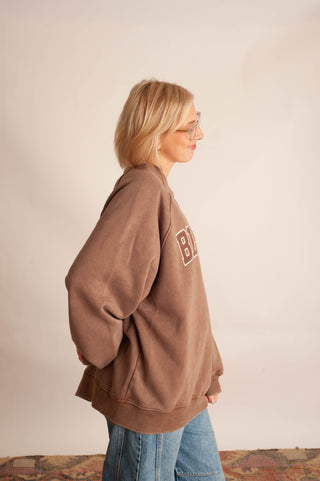 A woman with blonde hair and glasses is shown from the side profile, wearing an oversized brown sweatshirt with a white graphic block on the chest and blue jeans, standing against a plain beige wall.