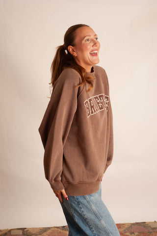 A woman wearing a brown sweatshirt with 'BICHES' printed in white across the chest, paired with blue jeans, posing against a plain beige wall.