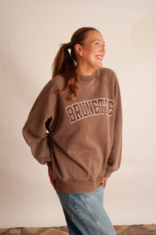 A woman wearing a brown crewneck sweatshirt with 'BRUNETTE' printed in arched varsity letters across the chest, paired with light blue jeans, standing against a plain beige wall.