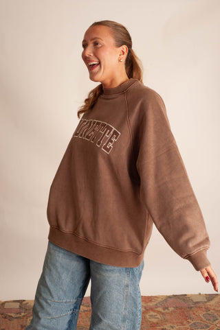 A woman wearing a brown crewneck sweatshirt with 'VIRGINIA' printed in arched varsity letters across the chest, paired with light blue jeans, standing against a plain beige wall.
