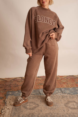 A person modeling a matching brown sweatshirt and jogger pants set with 'BLONDE' printed across the chest, standing on a patterned rug against a plain wall.
