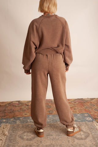 A person shown from the back wearing a matching brown sweatshirt and jogger pants set, standing on a patterned rug against a plain light wall.