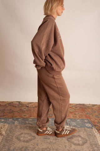 A woman wearing a matching brown lounge set consisting of a sweatshirt and jogger pants, standing on a patterned rug against a plain beige wall.