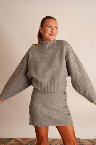 A woman modeling a heather gray knit two-piece set consisting of a cropped turtleneck sweater with balloon sleeves and a matching button-front mini skirt, set against a plain beige wall.