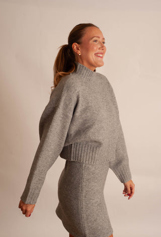 A woman wearing a heather gray knit mock-neck sweater and matching knit skirt, photographed against a plain light beige studio background.