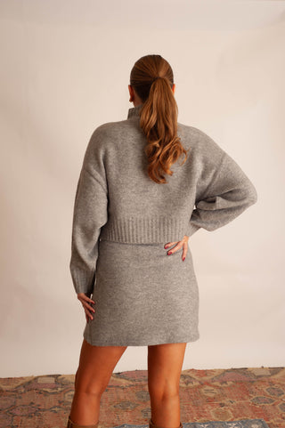A woman shown from the back wearing a light gray knit mock-neck sweater and matching knit skirt, standing against a plain beige wall with a patterned rug visible at the bottom.