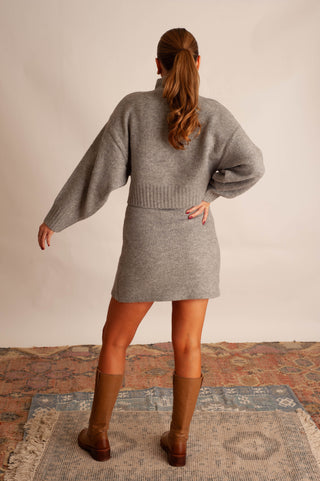 A woman viewed from behind wearing a heather gray knit sweater dress with balloon sleeves, paired with brown knee-high boots, standing on a patterned rug against a plain beige wall.