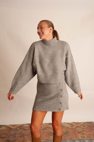 A woman modeling a light gray knit mock-neck sweater and matching button-front mini skirt set, standing against a plain beige wall with a patterned rug visible at the bottom.