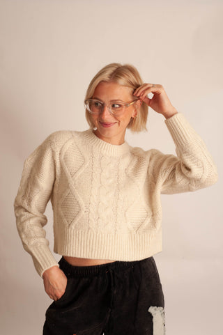 A woman with short blonde hair and glasses wearing a cream cable-knit cropped sweater and black pants, posing against a plain light beige studio background.