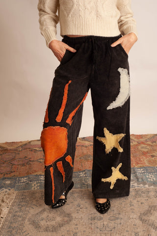 A person wearing wide-leg black pants with large orange sun and white moon patches, paired with a cream sweater and black pointed shoes, standing on a patterned rug.