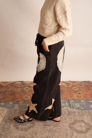 A person wearing a cream cable-knit sweater and a black wide-leg patchwork skirt with beige and gold starfish motifs, standing on a patterned rug against a neutral wall.