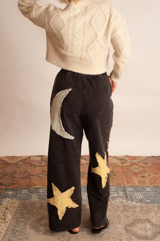 A person shown from the back wearing dark wide-leg pants with large cream moon and star appliqués, paired with a cream cable-knit cropped sweater, standing indoors on a patterned rug.