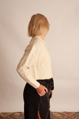 A woman with short blonde hair is shown from the back wearing a cream-colored cable-knit cropped sweater and black pants, standing against a plain light beige wall.