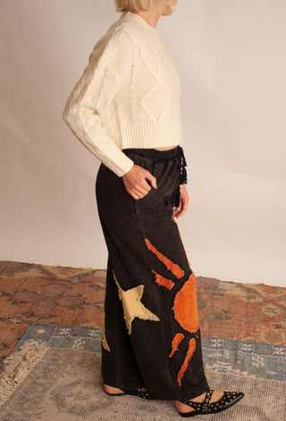 A model is shown from the side wearing a cream cable-knit cropped sweater and black wide-leg pants with orange and beige abstract patchwork details. The model is standing on a patterned rug against a plain beige wall.