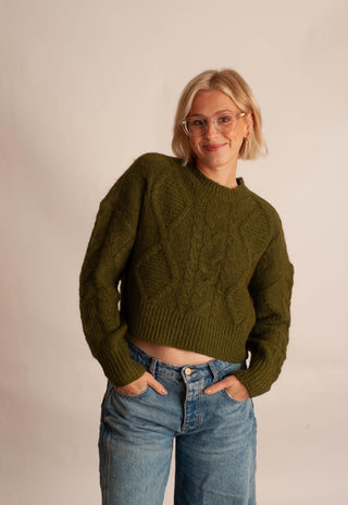 A woman with blonde hair and glasses wearing a cropped olive green cable-knit sweater and blue jeans, standing against a plain light beige background.