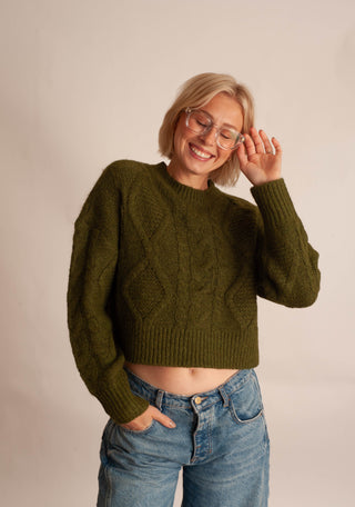 A woman with short blonde hair and glasses wearing an olive green cropped cable-knit sweater and blue jeans, posing against a plain beige studio background.