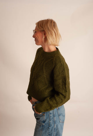 A woman with short blonde hair and glasses, wearing a textured olive green cable-knit sweater and blue jeans, standing in profile against a plain off-white background.