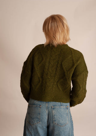 A person shown from the back wearing an olive green cable-knit cropped sweater and light blue jeans against a plain beige studio background.
