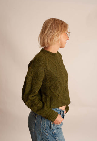 A woman with short blonde hair and glasses, wearing a textured olive green cable-knit sweater and blue jeans, shown from a side profile against a plain beige studio background.