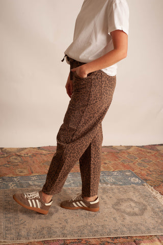 A person wearing brown leopard-print trousers, a white t-shirt, and brown and white sneakers, standing on a patterned rug against a plain beige wall.