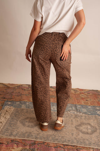 A person shown from the back wearing brown leopard-print pants, a white t-shirt, and brown platform sandals, standing on a patterned rug against a plain beige wall.