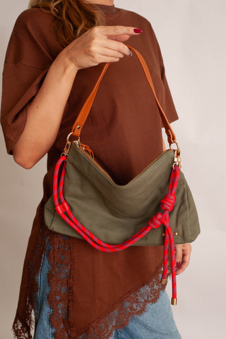 A woman wearing a brown t-shirt and blue jeans carries an olive green slouchy shoulder bag with a bright red braided rope detail and tan leather strap.