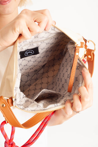 Close-up of a person holding open a beige shoulder bag to show the interior lining and gold-tone zipper. The bag has tan leather trim, a red-and-white patterned strap, and a black interior label with a white logo.